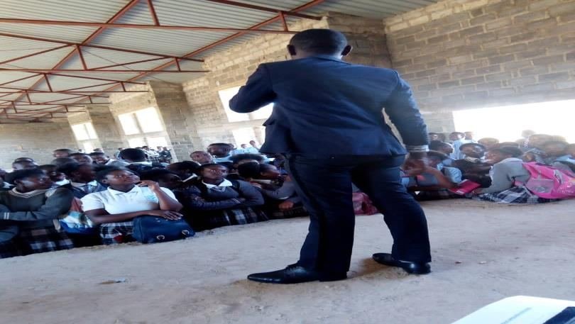 An MCUSzambia facilitator addressing a large group of seated secondary school pupils in an open hall
