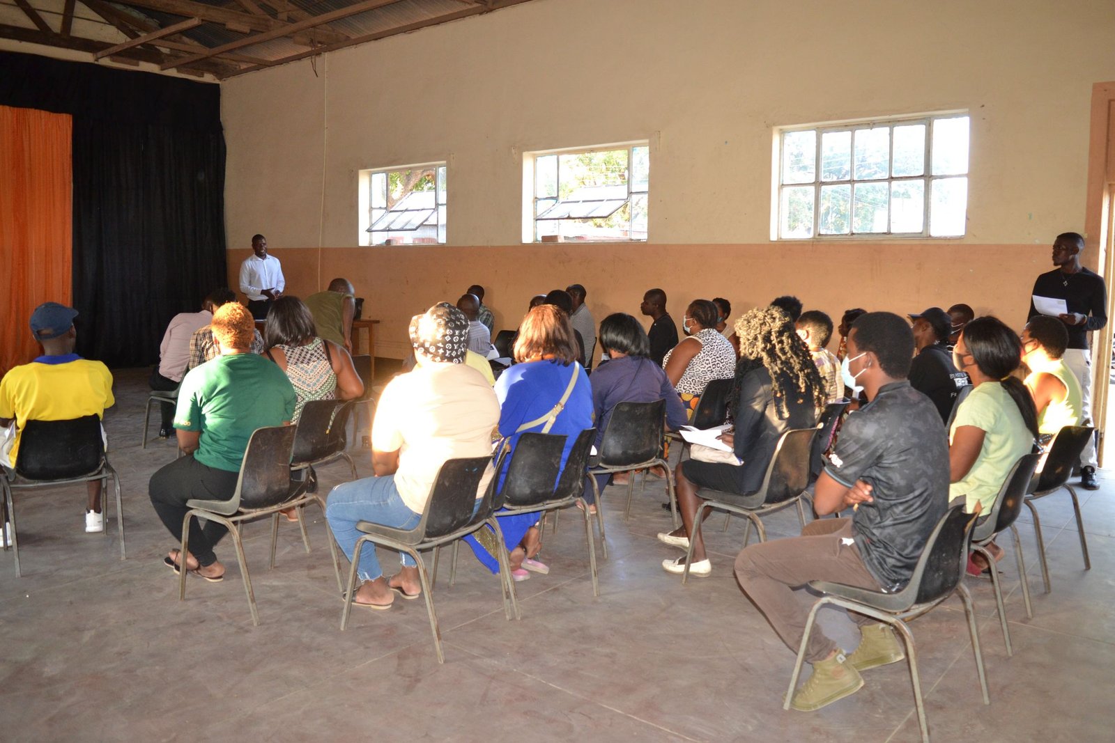 Adults seated on chairs in a school hall listening to a facilitator standing at the front