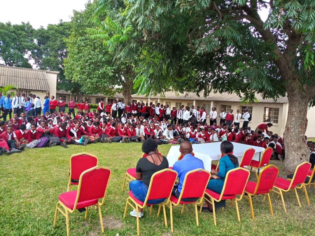 A large outdoor assembly of pupils in red and white uniform seated on the grass under trees