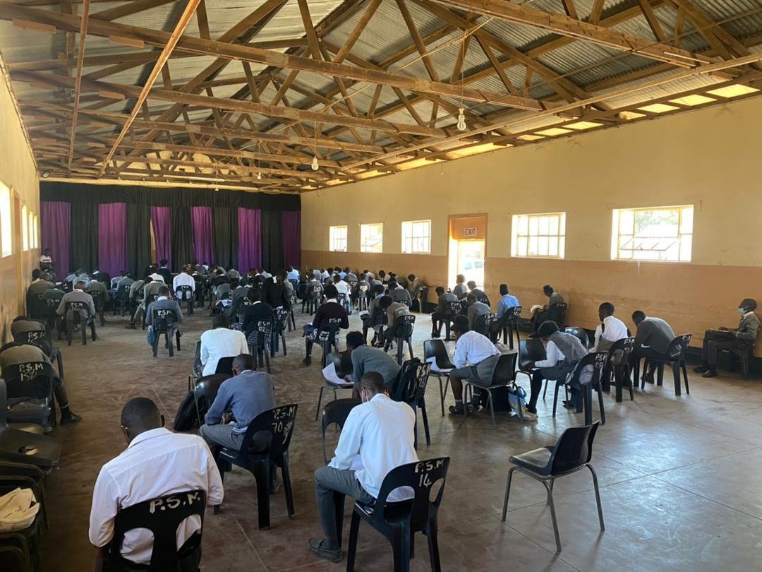 Pupils seated at desks across a large school hall with a corrugated iron roof