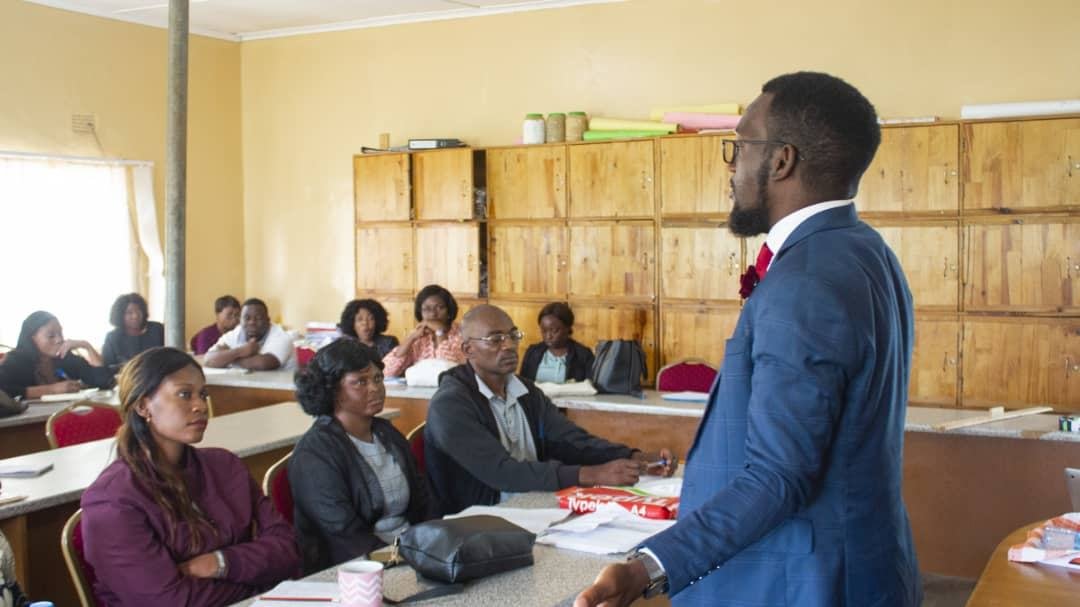 A facilitator in a blue suit standing and speaking to a group seated at desks