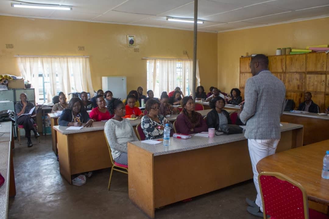 Adults seated at desks in a classroom while a facilitator stands at the front of the room