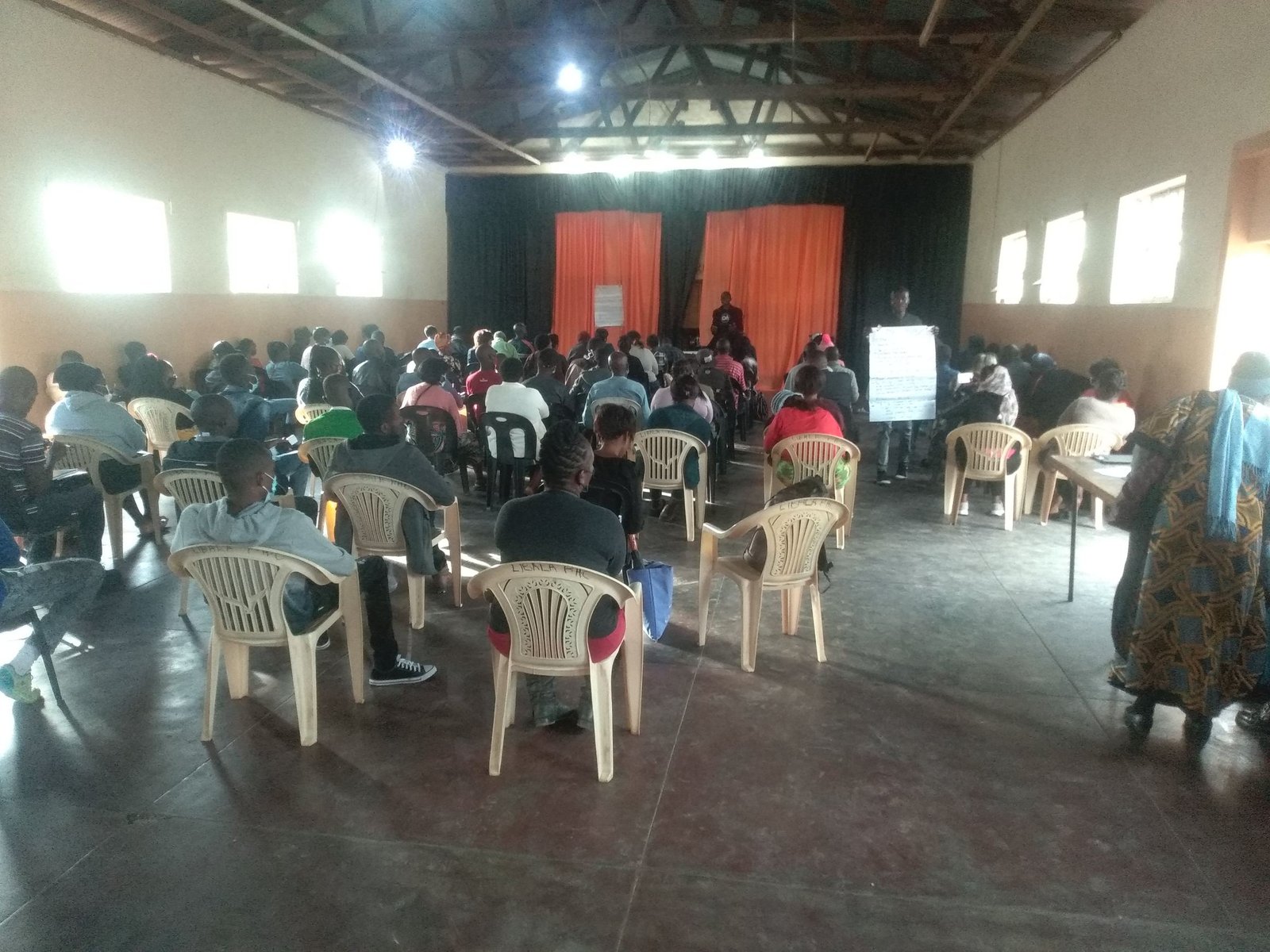 A hall of people seated on plastic chairs facing a facilitator holding a flipchart