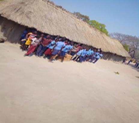 Pupils in blue uniforms sitting in the shade under the low thatched eaves of the school building