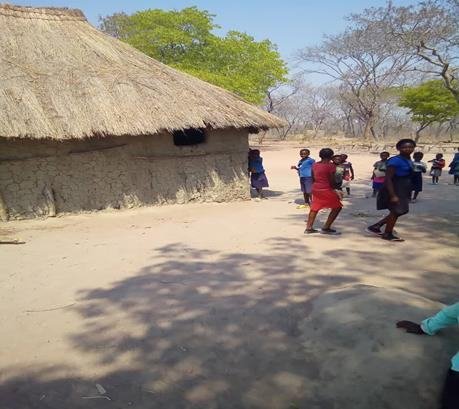 Children walking across the open sand yard between thatched buildings at Nanungwe Primary School