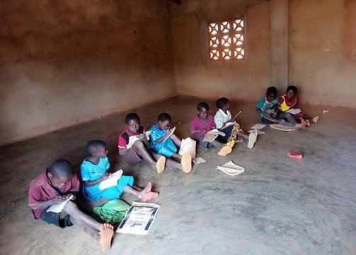 Young pupils sitting on the bare floor of a classroom, working from exercise books